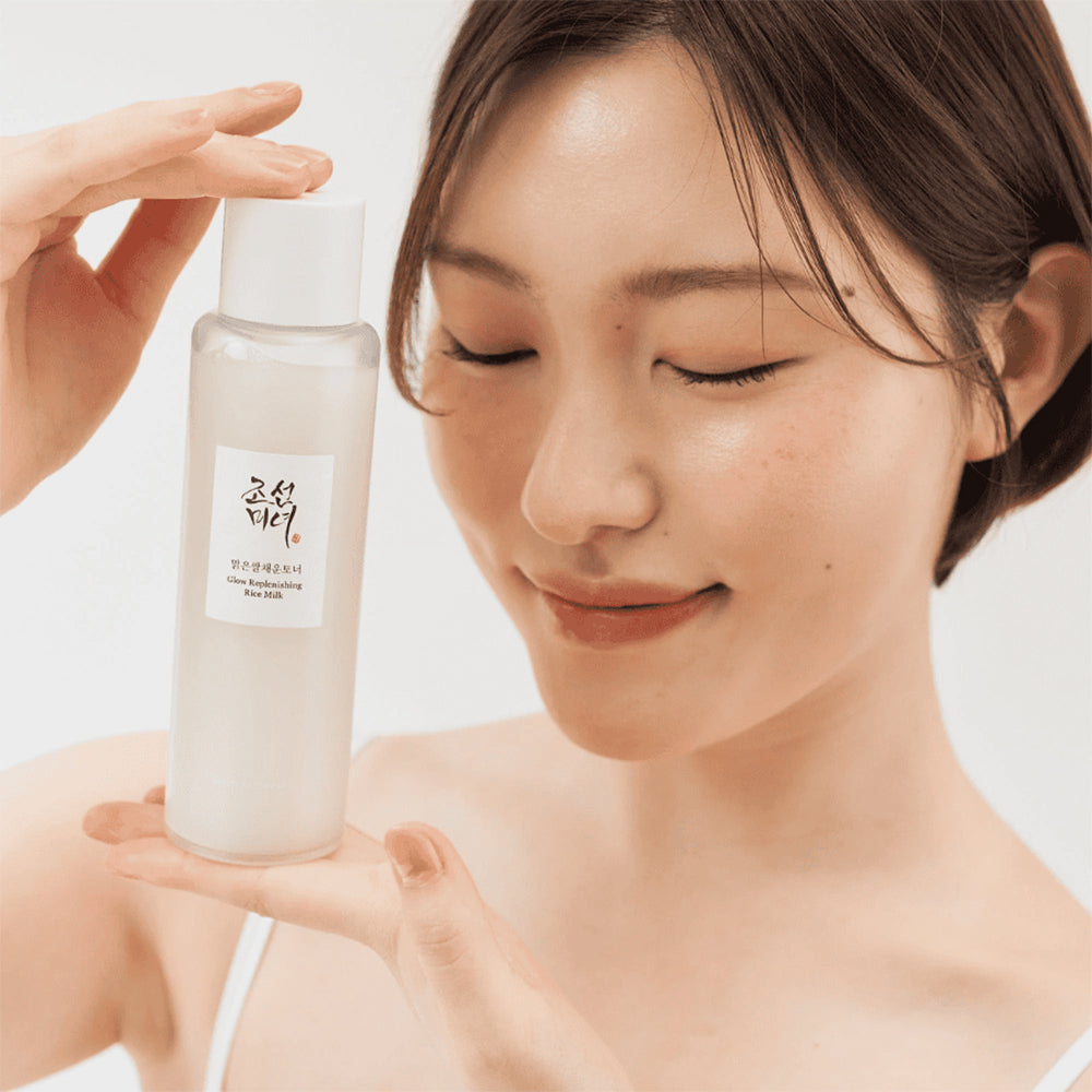 Kvinde med glødende hud holder Beauty Of Joseon Glow Replenishing Rice Milk 150ml foran ansigtet og smiler med lukkede øjne.