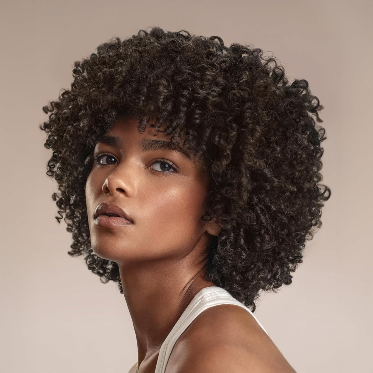 Kvinde med naturligt krøllet afro-hår og curly bangs, iført hvid top mod beige baggrund.