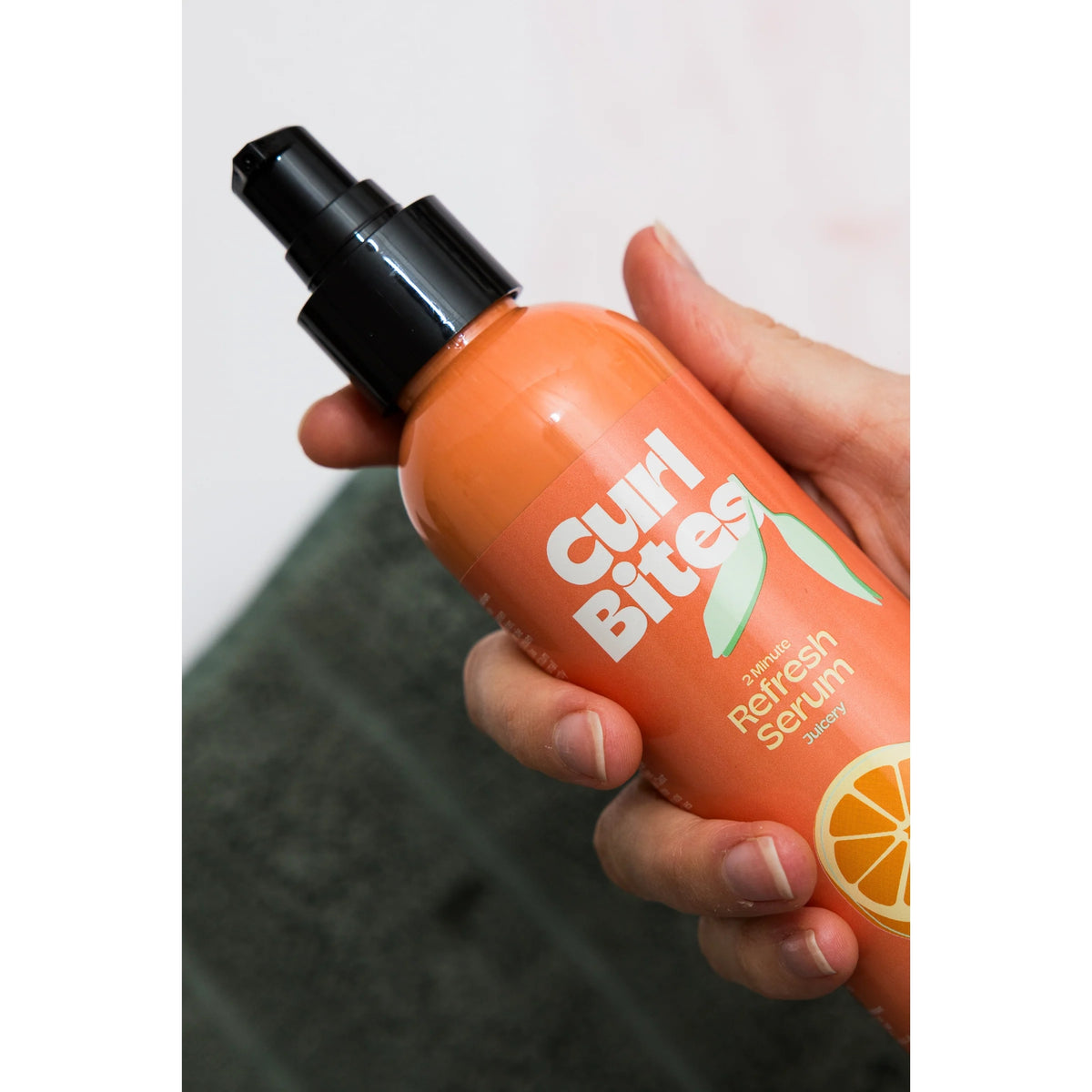 Hånd holder orange Curl Bites 2 Minute Refresh Serum flaske med sort pumpe mod grøn baggrund.