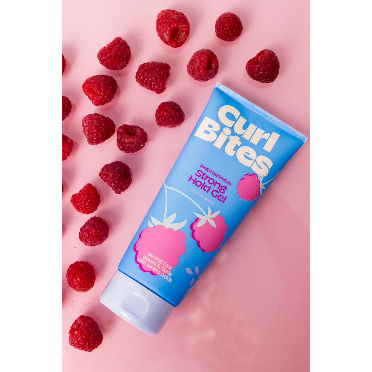 Curl Bites Strong Hold Gel i blå tube med hindbær, omgivet af friske hindbær på lyserød baggrund.