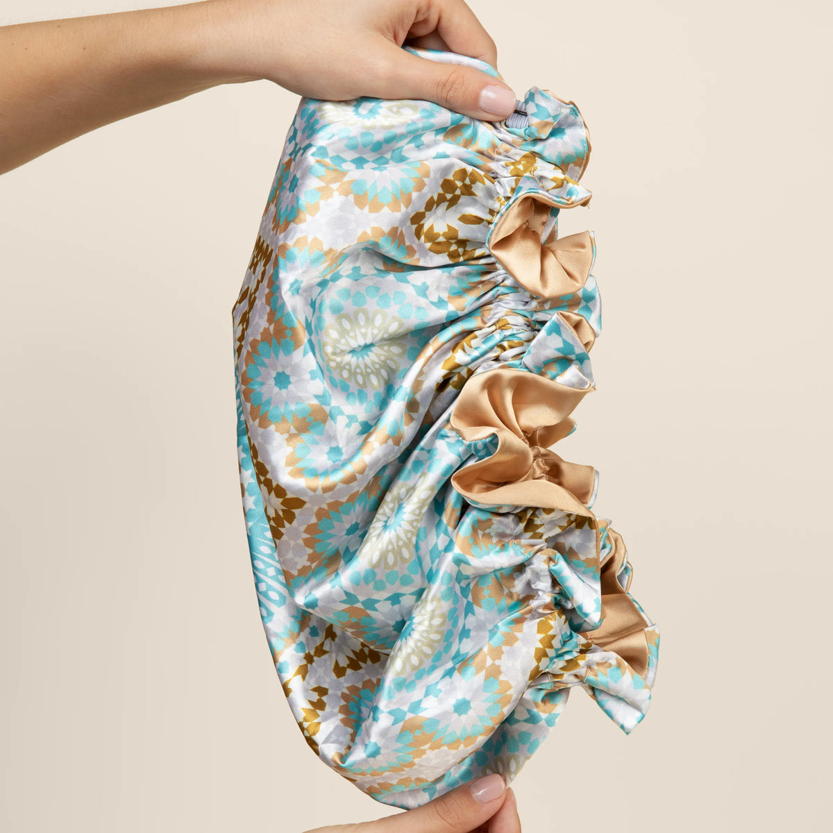 Bounce Curl Reversible Satin Hair Bonnet i mintgrøn, guld og hvidt mønster holdes op mellem to hænder.